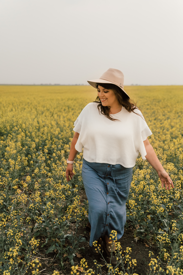 Dani wearing a white top, a long denim skirt and a beige hat, walks through a field