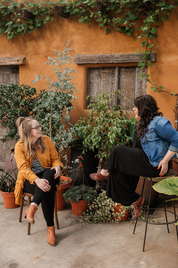 two women sit outside with plants and a house behind them