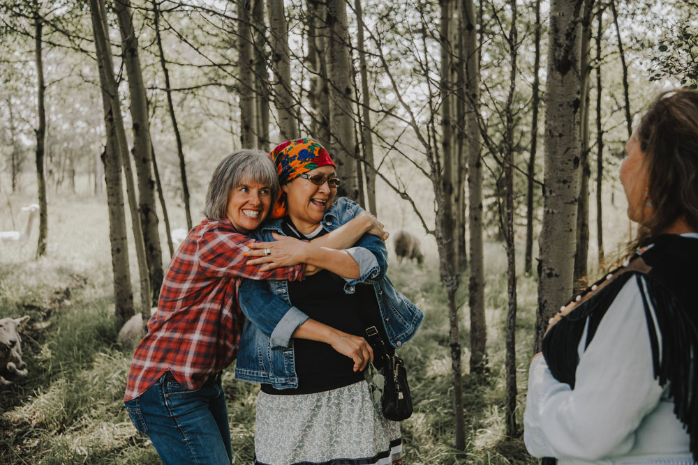 Krista and Tara embrace as Alanna looks on, they are surrounded by trees